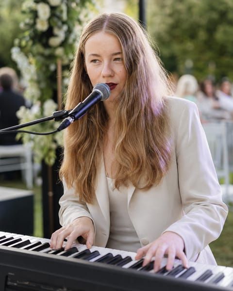 Jess Martyn singing at the keyboard at an outdoor garden wedding, floral arch and guests behind