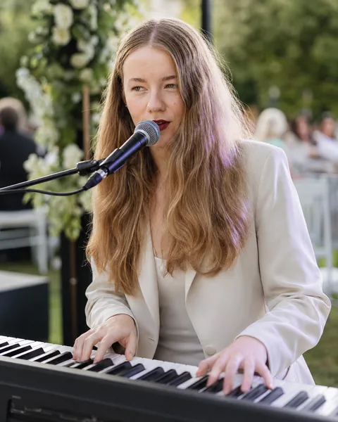Jess Martyn singing at the keyboard at an outdoor garden wedding, floral arch and guests behind