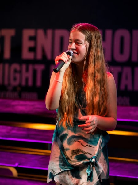 Jess Martyn mid-song with a handheld microphone, magenta and orange stage lighting