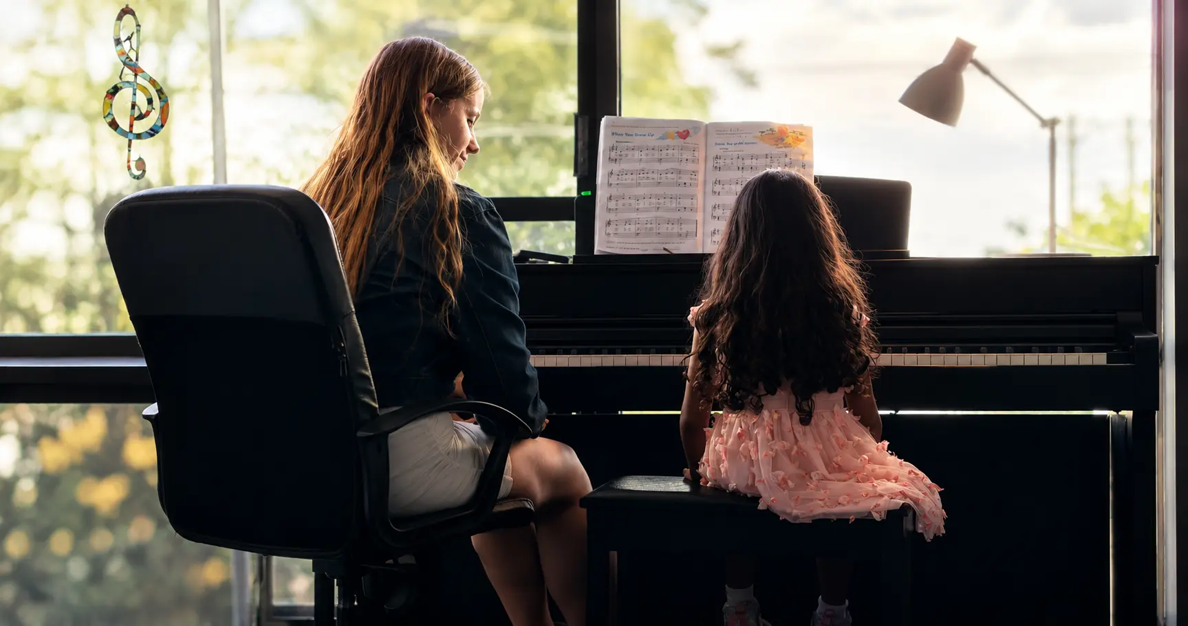 Jess teaching a young student at an upright piano beside a bright window, sheet music open in front of them